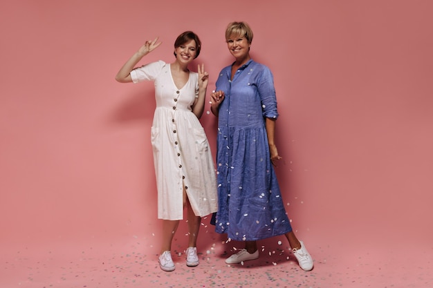 Full length photo of girl with short hair in white dress and sneakers smiling, showing peace sign and posing with blonde woman on pink backdrop. 