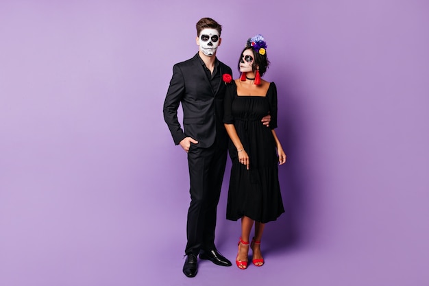 Full-length photo of guy and girl in elegant black outfit and halloween masks posing on purple wall.