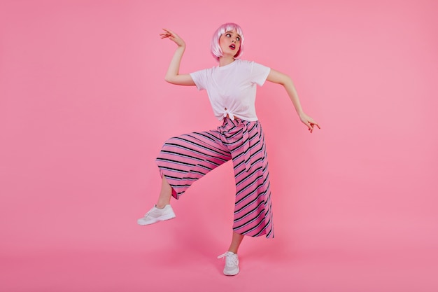 Full-length photo of joyful girl in peruke fooling around during . Magnificent funny lady in periwig dancing on pink wall