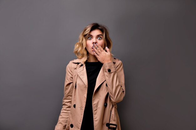 Full-length photo of joyful girl with short hairstyle in beige casual trench covering mouth by hand. Magnificent funny lady standing over isolated grey wall