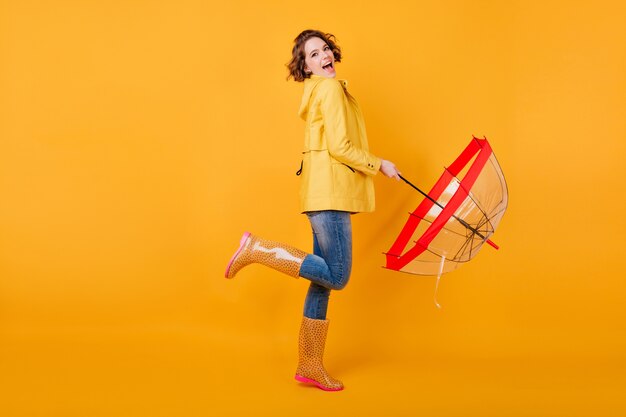 Фото в полный рост довольной девушки в модной осенней куртке, стоящей на одной ноге. Возбужденная европейская женская модель с зонтиком, выражающим положительные эмоции на желтой стене.