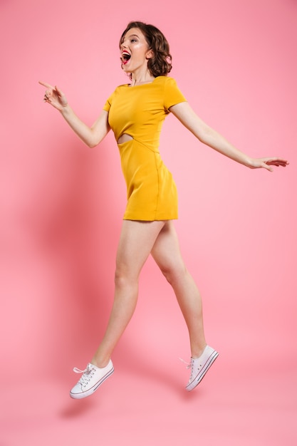 Full length photo of pretty girl with red lips makeup pointing with finger, looking aside while jumping over pink 