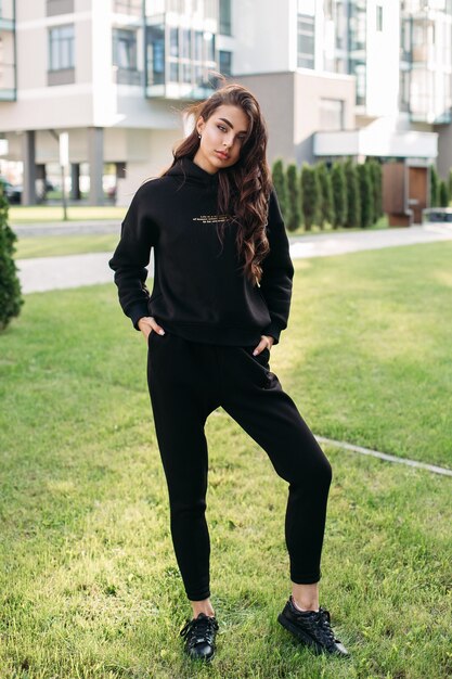 Фото в полный рост довольно молодой женщины в черной спортивной одежде, позирующей на улице, стоя на лужайке на открытом воздухе. Женская мода. Городской образ жизни