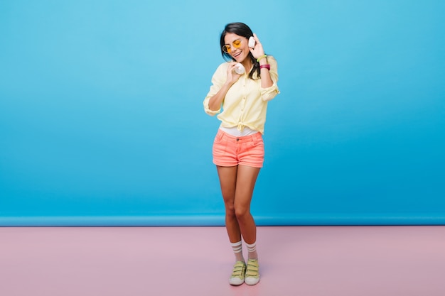 Full-length photo of shapely tanned girl in pink shorts dancing with pleasure. Catching dark-haired european woman in yellow shoes listening music in white earphones.