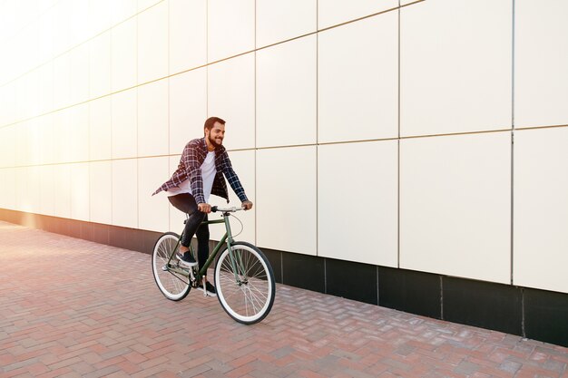 Full length photo of smiling young bearded man riding a bike on the city street.