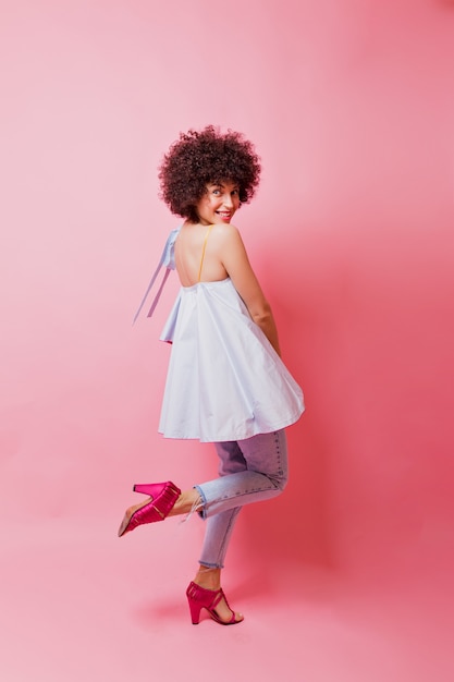 Free photo full-length photo of stylish charming woman with short curly hair dressed blue shirt, jeans and pink heels on pink