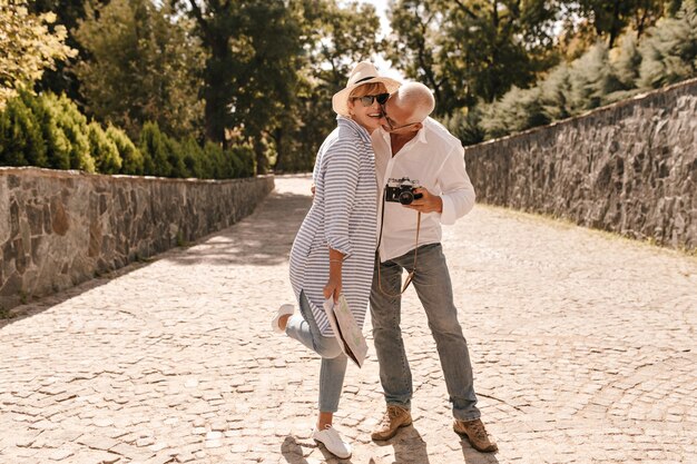 Full length photo of stylish woman in hat, sunglasses and long blue blouse laughing with grey haired man in white shirt and jeans with camera outdoor.