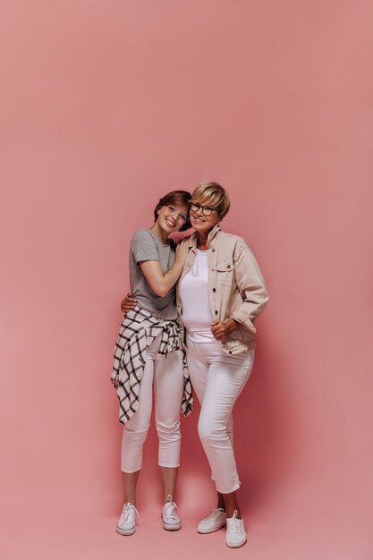 짧은 멋진 헤어 스타일과 흰 바지와 운동화에 현대적인 안경을 쓰고 웃고 고립 된 배경에서 포옹하는 두 여성의 전체 길이 사진.