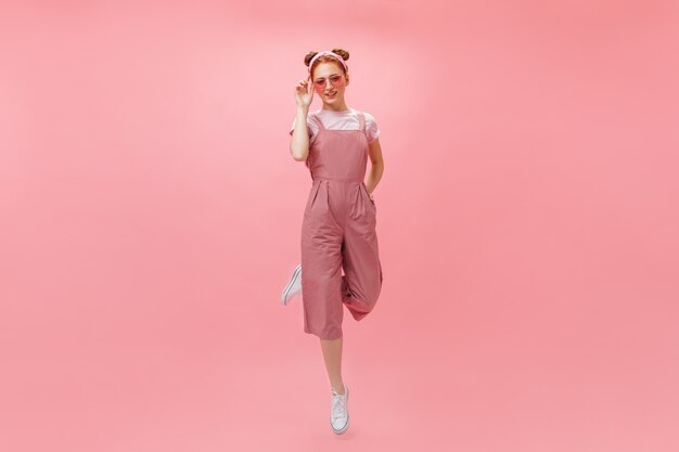 Full-length photo of woman in pink jumpsuit and accessories jumping on isolated background.