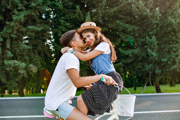 Фото молодой пары в полный рост на велосипеде на дороге на фоне леса. Парень в белой футболке едет на велосипеде и целует девушку, сидящую на руле