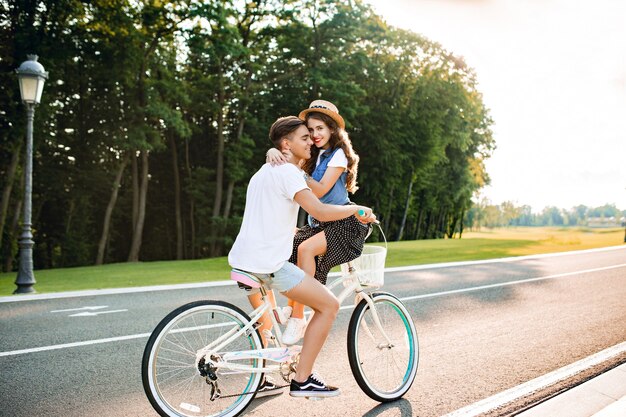 Full-length photo of young couple in love on bike on road. A guy in white T-shirt is driving a bike and kissing a girl sitting on the handlebar