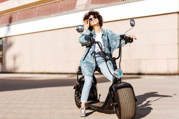 現代のバイクの屋外に座っているとよそ見サングラスの屈託のない巻き毛の女性の完全な長さの画像