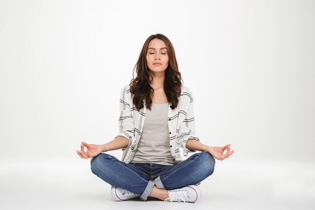 Full-length picture of concentrated woman in casual clothes meditating with closed eyes while sitting in lotus pose on the floor, isolated over white wall