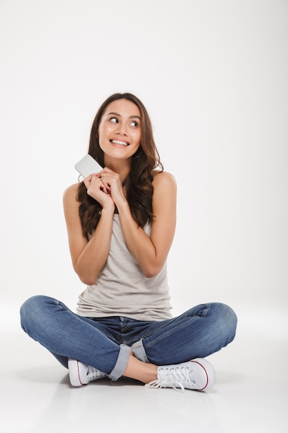 Full-length picture of happy woman in jeans and sneakers holding modern cellphone while sitting with legs crossed on the floor, over white wall