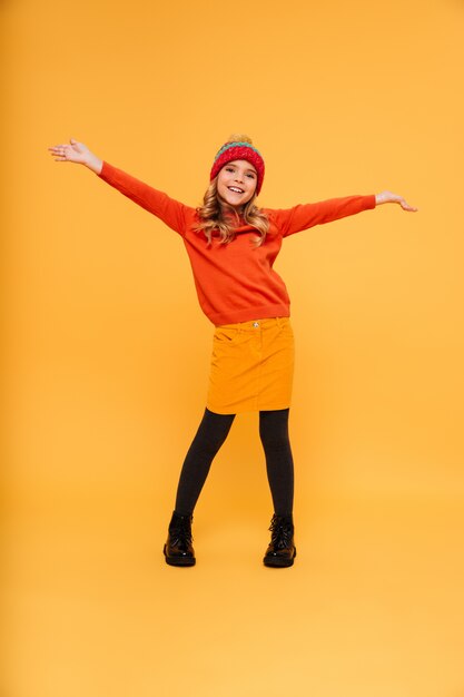 セーターと帽子のポーズとオレンジの上のカメラを見て完全な長さの遊び心のある若い女の子