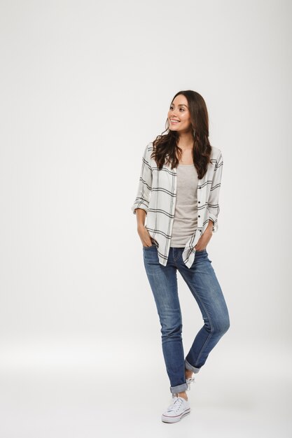 Full length Pleased brunette woman in shirt posing with arms in pockets and looking away over gray