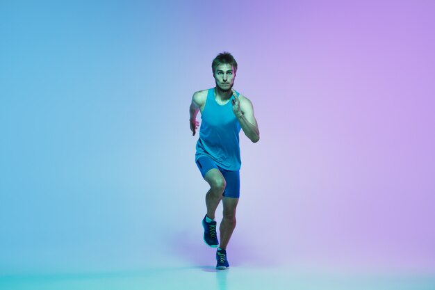 Full length portrait of active young caucasian running, jogging man on gradient studio in neon light