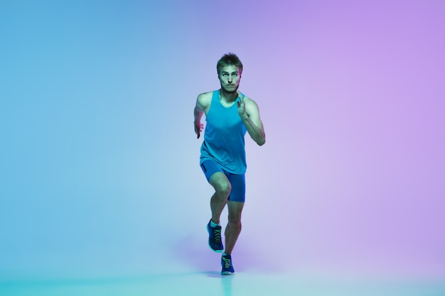 Full length portrait of active young caucasian running, jogging man on gradient studio in neon light