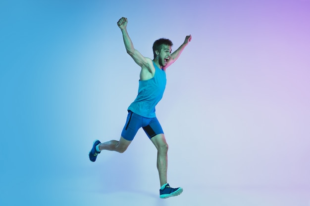 Full length portrait of active young caucasian running, jogging man on gradient studio in neon light