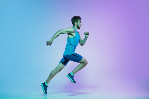 Full length portrait of active young caucasian running, jogging man on gradient studio in neon light