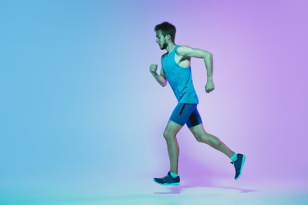 Full length portrait of active young caucasian running, jogging man on gradient studio in neon light