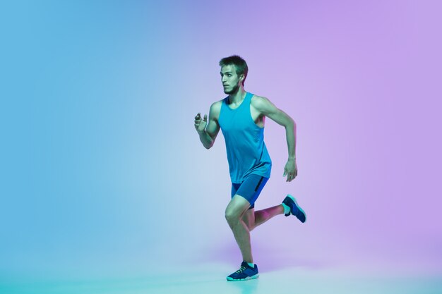 Full length portrait of active young caucasian running, jogging man on gradient studio in neon light