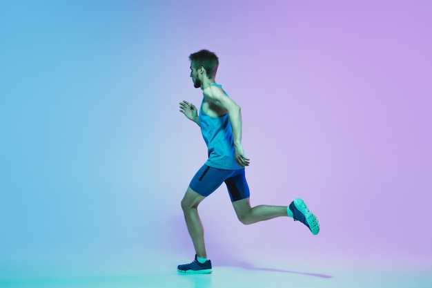 Free photo full length portrait of active young caucasian running, jogging man on gradient studio in neon light