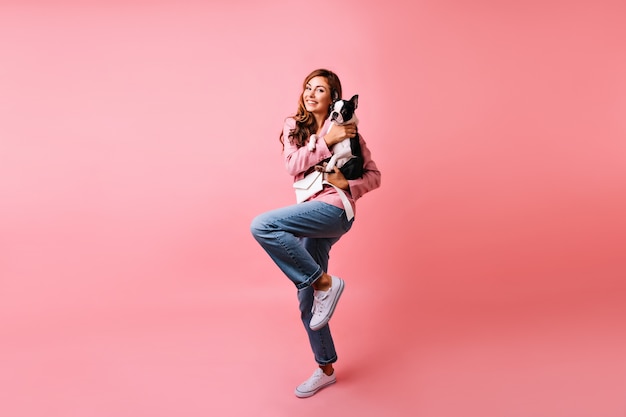 Full-length portrait of adorable girl in trendy jeans holding french bulldog. Dreamy caucasian woman posing with her dog on pink.