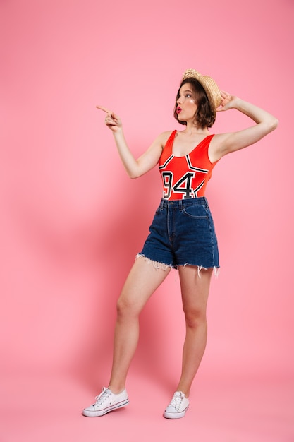 Full length portrait of amazed young woman in red bodysuit and jeans short holding her straw hat, pointing with finger, looking aside