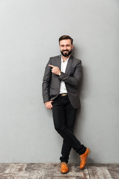 Full-length portrait of attractive man posing on camera with broad smile pointing index finger aside, isolated over gray