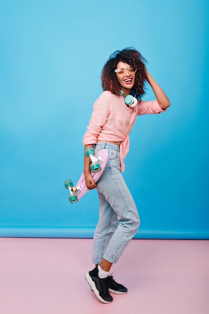 Full-length portrait of attractive sporty girl with brown skin dancing with longboard.