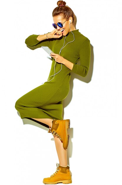 헤드폰으로 스마트 폰에서 흰색 듣는 음악에 고립 된 캐주얼 녹색 hipster 여름 옷에서 아름 다운 행복 귀여운 웃는 갈색 머리 여자 여자의 전체 길이 초상화