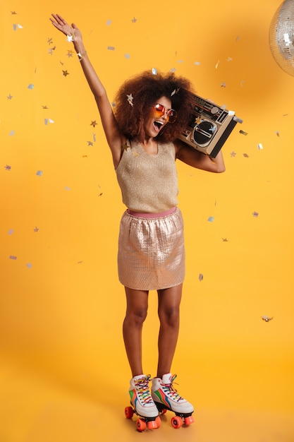 Foto gratuita ritratto integrale di bella donna afroamericana felicissima della discoteca con la mano sollevata, stando sui pattini di rullo, tenendo boombox