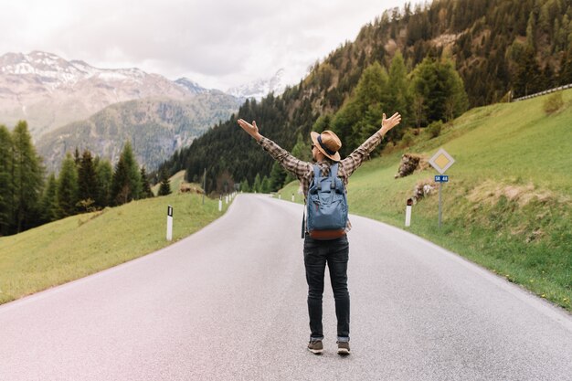 Full-length portrait of blissful man with backpack emotionally posing with hands up and hitchhiking on the highway