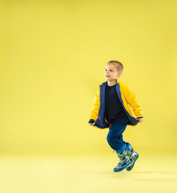 Портрет в полный рост яркого модного мальчика в плаще, бегущего и веселого на желтом.