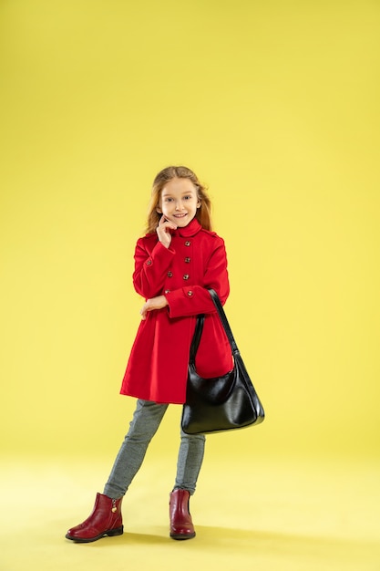 A full length portrait of a bright fashionable caucasian girl in a red raincoat holding a black bag on yellow.