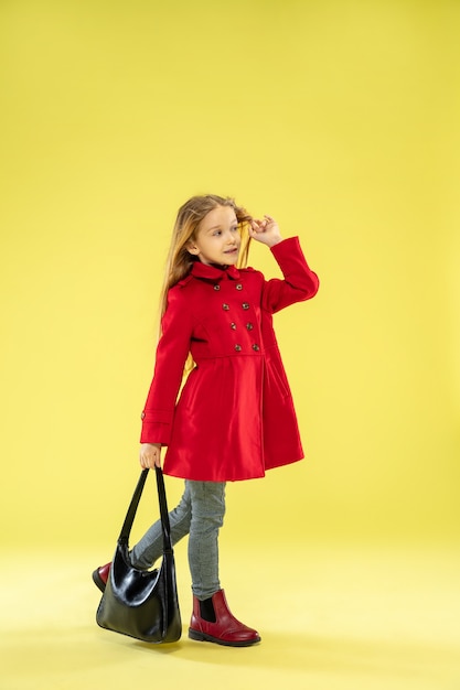 A full length portrait of a bright fashionable caucasian girl in a red raincoat holding a black bag on yellow