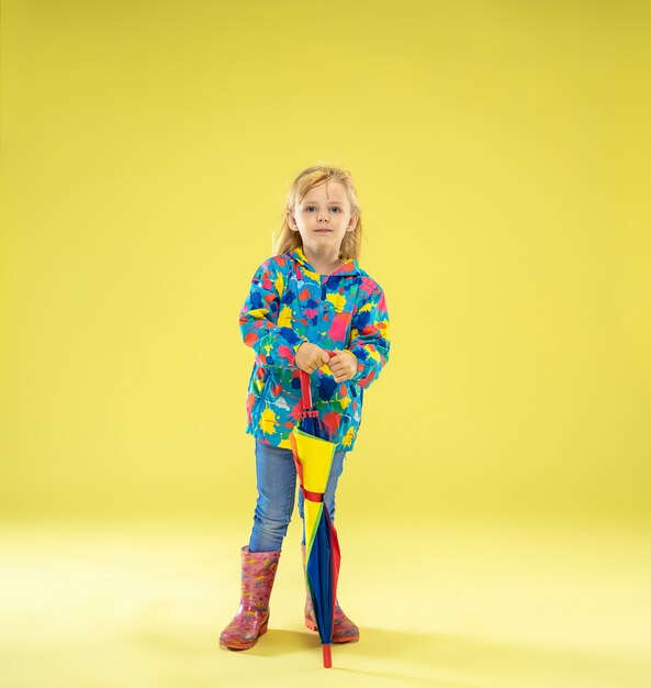 A full length portrait of a bright fashionable girl in a raincoat holding an umbrella of rainbow colors on yellow studio wall