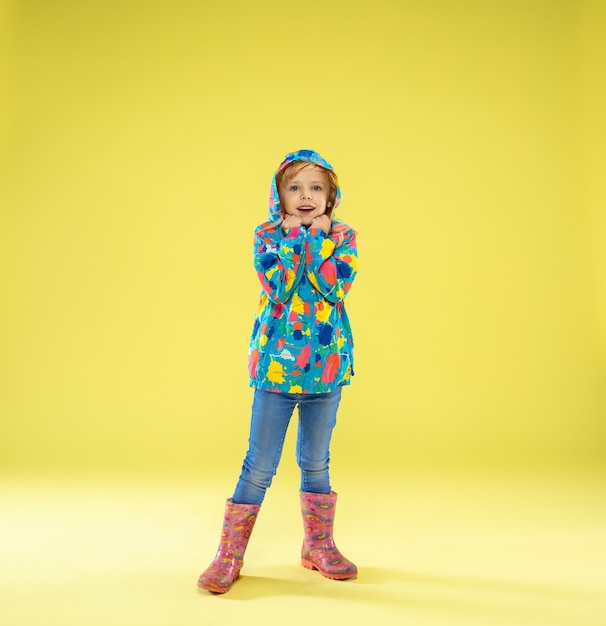 A full length portrait of a bright fashionable girl in a raincoat holding an umbrella of rainbow colors on yellow.