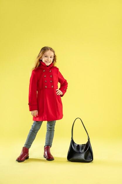 Портрет яркой модной девушки в красном плаще с черной сумкой в полный рост, позирующей на желтом студийном фоне.