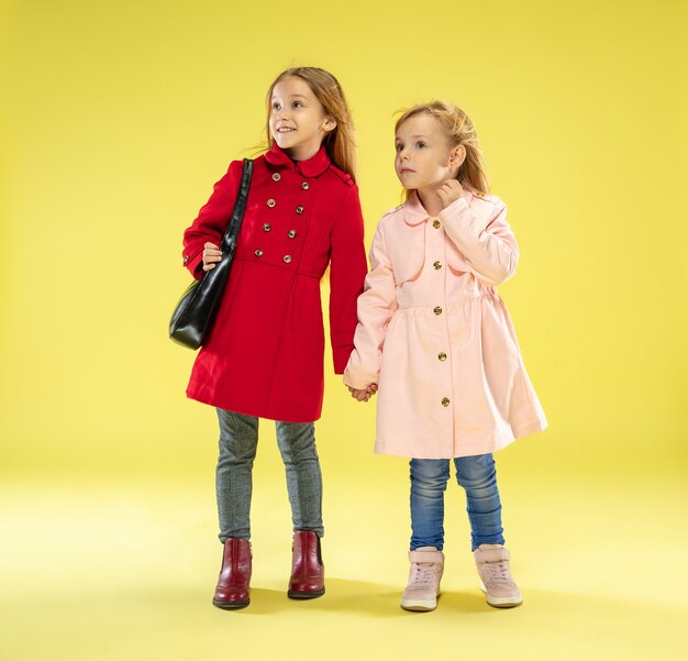 A full length portrait of a bright fashionable girls in a raincoat holding a black bag on yellow studio wall