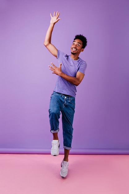 Free photo full-length portrait of brunette man in trendy blue jeans. african male model in stylish purple t-shirt standing.