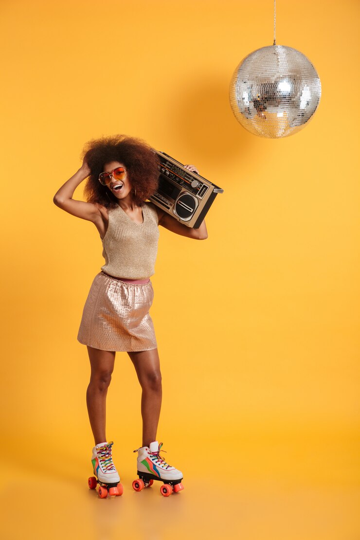 Free Photo full length portrait of charming african woman in retro clothes standing on roller skates, holding boombox, touching her afro hairstyle