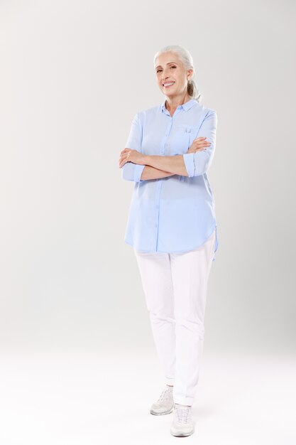 Full-length portrait of charming old lady in blue shirt and white pants, standing with crossed hands