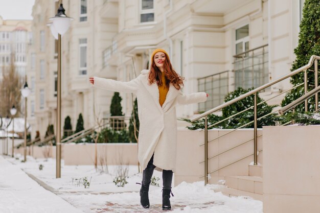 Full-length portrait of cheerful caucasian woman smiling on snowy street. Merry ginger girl having fun in cold day.