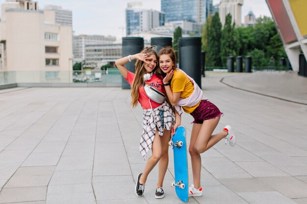 Full-length portrait of cheerful funny girl stading on one leg with blue skateboard beside best friend