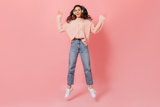 Full-length portrait of cheerful lady jumping on pink background. Stylish girl in headphones and knitted sweater listens to music.