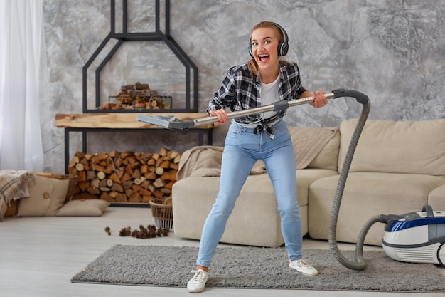 Full length portrait of cheerful young woman 20s listening to music via headphones and having fun with vacuum cleaner in the house. Busy, cleaning day. Home, housekeeping concept