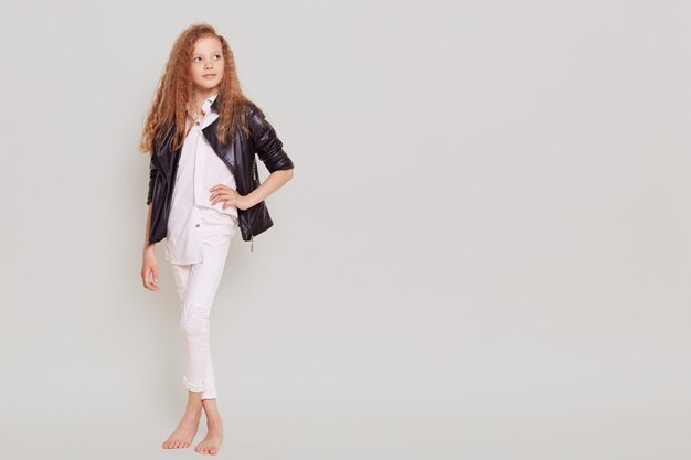 Full length portrait of confident little schoolgirl wearing white clothing and leather jacket looking away and keeping hand on hip