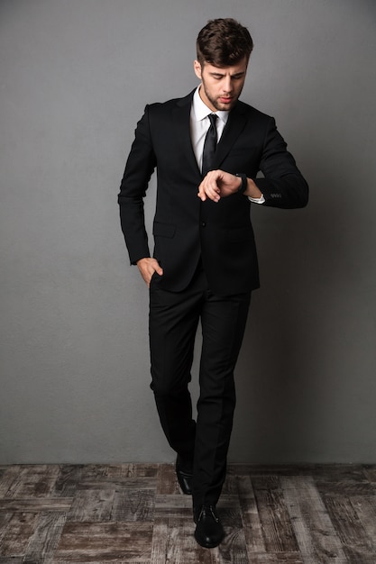 Full length portrait of confident man in classic black suit checking time on his wristwatch